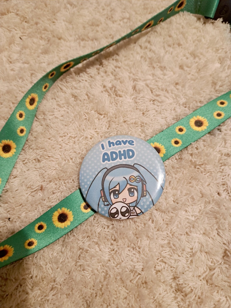 May include: A light blue button with the text "I have ADHD" and a cartoon character illustration. The button is on a green lanyard decorated with yellow sunflowers. The lanyard is on a white, textured surface.