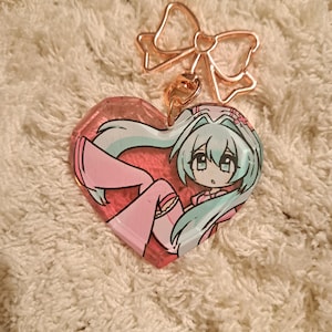 May include: A heart-shaped keyring featuring a rose gold bow-shaped clasp and a translucent pink resin charm. The charm displays an anime-style character with long teal hair, wearing pink clothing, set against a textured, light-coloured background.