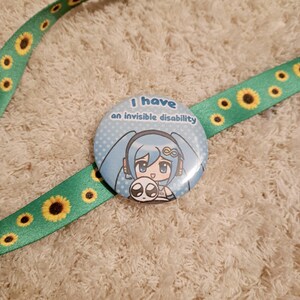 May include: A round button with the text "I have an invisible disability" and a cartoon character with blue hair and a small creature. The button is on a green lanyard with yellow sunflower designs.