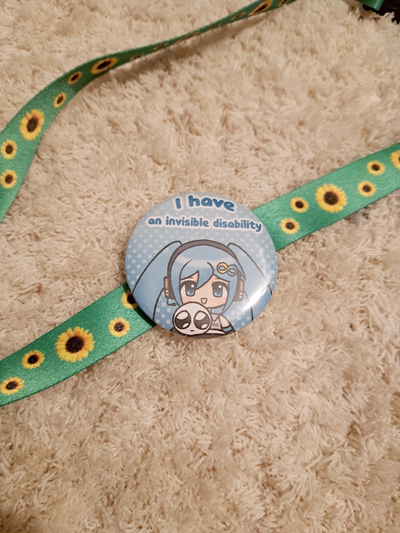 May include: A round button with the text "I have an invisible disability" and a cartoon character with blue hair and a small creature. The button is on a green lanyard with yellow sunflower designs.