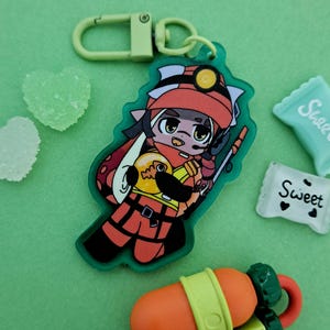 May include: A green and orange cartoon character keychain with a red hat and a fish in its hand. The keychain has a green and silver metal ring.