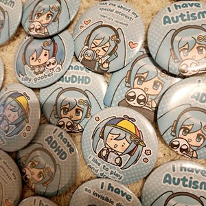 May include: A selection of light blue pin badges with anime-style illustrations. Some badges display text such as "I have Autism" and "I have ADHD." Other badges have phrases like "ask me about my special interest!" and "I like to play and draw."