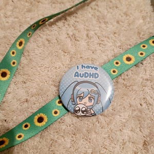 May include: A green lanyard with yellow sunflowers and a round button. The button has a blue background with a cartoon character and the text "I have AUDHD". The lanyard is on a cream-colored, textured surface.