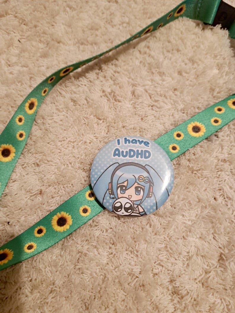 May include: A green lanyard with yellow sunflowers and a round button. The button has a blue background with a cartoon character and the text "I have AUDHD". The lanyard is on a cream-colored, textured surface.