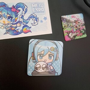 May include: A collection of colorful anime-style merchandise. Includes a square button featuring a blue-haired character with headphones and a small creature, a sticker with a mermaid-like character, and a Splatoon 2 button.