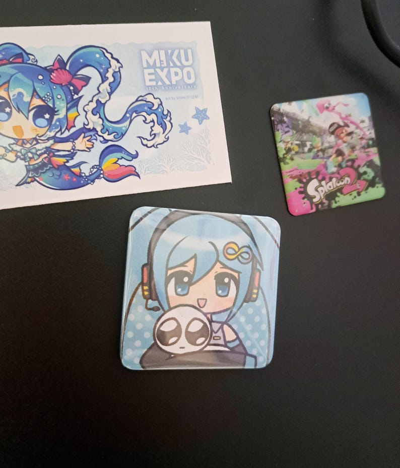 May include: A collection of colorful anime-style merchandise. Includes a square button featuring a blue-haired character with headphones and a small creature, a sticker with a mermaid-like character, and a Splatoon 2 button.