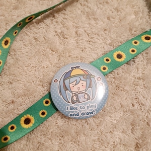 May include: A round, light blue button with a cartoon character and the text "I like to play and draw!" is attached to a green ribbon decorated with yellow sunflowers. The ribbon is lying on a white, textured surface.