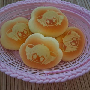 Könnte beinhalten: Ein rosa Plastikkorb enthält vier wolkenförmige Gebäckstücke. Jedes Gebäckstück hat einen gelb-orangen Farbverlauf und ein Cartoon-Engelgesicht-Design. Der Korb steht auf einer hellbraunen Oberfläche.