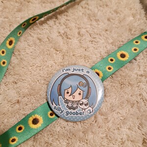 May include: A round button with a cartoon character and the text "I'm just a silly goober :3" is attached to a green lanyard with yellow sunflowers. The button features a blue-haired anime-style character with headphones and a small white creature.