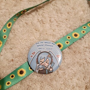 May include: A round, light blue button with an anime-style illustration of a character with long, light blue hair. The button has the text "ask me about my special interest! (HINT-it's vocaloid!)" and is on a green lanyard with yellow sunflowers.