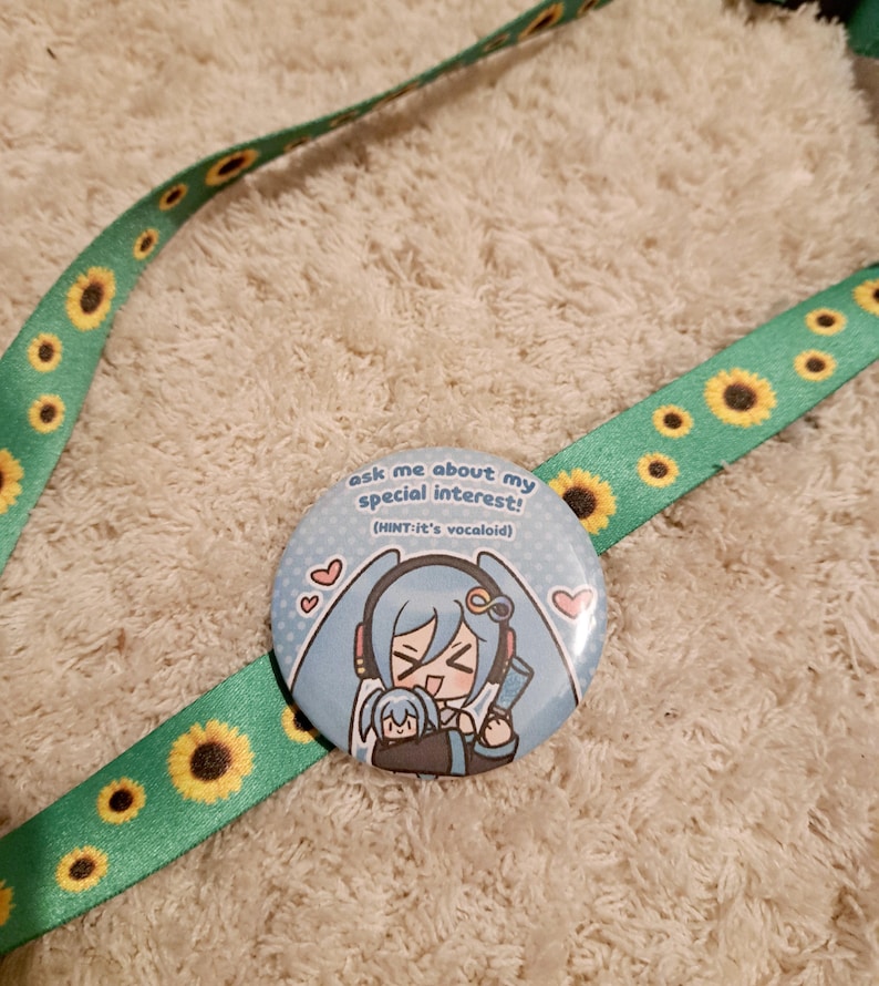 May include: A round, light blue button with an anime-style illustration of a character with long, light blue hair. The button has the text "ask me about my special interest! (HINT-it's vocaloid!)" and is on a green lanyard with yellow sunflowers.