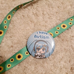 May include: A round button with the text "I have Autism" and a cartoon character with blue hair. The button is attached to a green lanyard decorated with yellow sunflowers.
