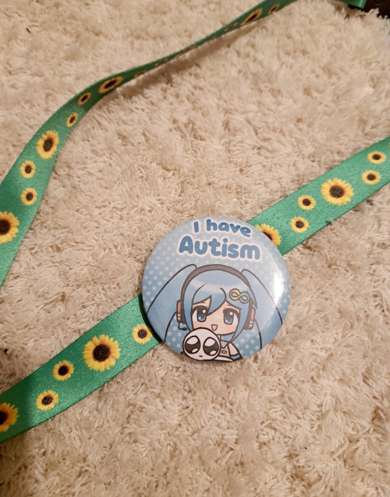 May include: A round button with the text "I have Autism" and a cartoon character with blue hair. The button is attached to a green lanyard decorated with yellow sunflowers.