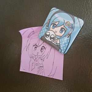 May include: A square, light blue button featuring an anime-style character with blue hair and headphones, holding a small, white skull. A pink sticky note with a pencil sketch of a similar character is next to the button.