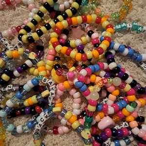 May include: A selection of vibrant beaded bracelets in a variety of colours, including yellow, orange, pink, blue, and black. Some bracelets have letter beads spelling out names and phrases. The bracelets are made of plastic beads.