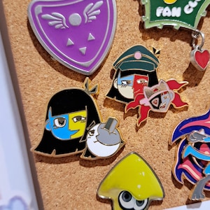 Puede incluir: Una colección de pines esmaltados con varios diseños de personajes de dibujos animados. Los pines presentan colores vibrantes, incluyendo morado, amarillo, rojo y azul, con contornos negros. Algunos pines tienen texto, y los pines están adheridos a un corcho.