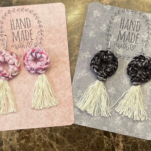 May include: Four pairs of handmade crocheted earrings with white tassels. The earrings are displayed on cards with the text "Hand Made with Love". The earrings are in four different colors: pink, black, blue, and a mix of pink and blue.