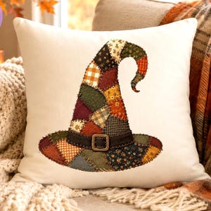 May include: A square white throw pillow featuring a central graphic of a witch hat constructed from a patchwork of various autumn-colored fabrics, including orange, brown, green, and yellow patterns, with a dark brown band and a bronze-colored buckle.