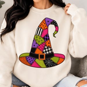 May include: A cream-colored crewneck sweatshirt featuring a large, central graphic of a witch hat. The hat is designed with a colorful patchwork pattern in shades of purple, orange, lime green, and black, including polka dots, stripes, and spiderwebs.