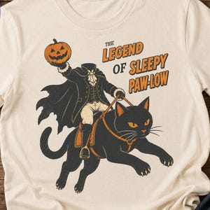 May include: A cream-colored t-shirt featuring a graphic of a headless figure in a black cape riding a large black cat while holding a jack-o'-lantern. The text reads THE LEGEND OF SLEEPY PAW-LOW in orange and black lettering on the front of the shirt.