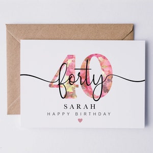 May include: A white card with a floral design in pink and yellow. The number 40 is written in black with the word "forty" written in cursive. The text "Sarah Happy Birthday" is written below the number. A small pink heart is at the bottom of the card.