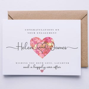 Puede incluir: Una tarjeta blanca con un coraz&oacute;n floral rosa y el texto "Felicidades por su compromiso Helen y James Les deseamos mucho amor, risas y un feliz para siempre".