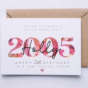May include: A white birthday card with the name "Holly" in black script, set against a floral design within the numbers "2005". The card reads "Happy 21st Birthday" and is placed on a brown envelope.