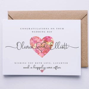 Op de afbeelding: Een witte trouwkaart met een roze bloemig hartontwerp. De kaart luidt "Gefeliciteerd met jullie huwelijk Olivia en Elliott We wensen jullie veel liefde, gelach en een gelukkig voor altijd".