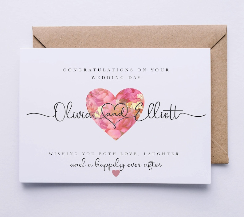 Op de afbeelding: Een witte trouwkaart met een roze bloemig hartontwerp. De kaart luidt "Gefeliciteerd met jullie huwelijk Olivia en Elliott We wensen jullie veel liefde, gelach en een gelukkig voor altijd".