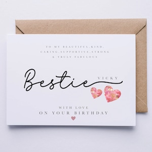May include: A white greeting card with the word "Bestie" in cursive script, accompanied by two pink heart illustrations. The card includes text that reads "To my beautiful, kind, caring, supportive, strong & truly fabulous" and "With love on your birthday."