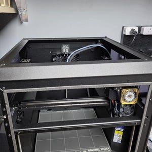 May include: A black Original Prusa 3D printer with a rectangular frame. The printer has a print bed, a nozzle assembly with a fan, and a control panel. The text "INPUT SHAPER" is visible on a component inside the printer.