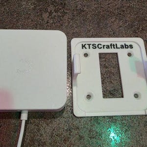 May include: A white electronic device and a white mounting bracket. The device has a rounded square shape with a small logo. The bracket has a rectangular opening and the text "KTSCraftLabs".