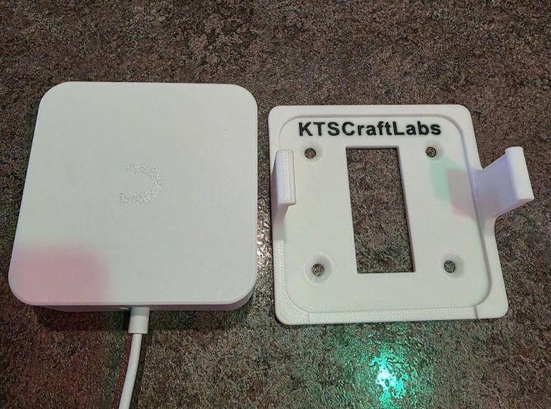May include: A white electronic device and a white mounting bracket. The device has a rounded square shape with a small logo. The bracket has a rectangular opening and the text "KTSCraftLabs".