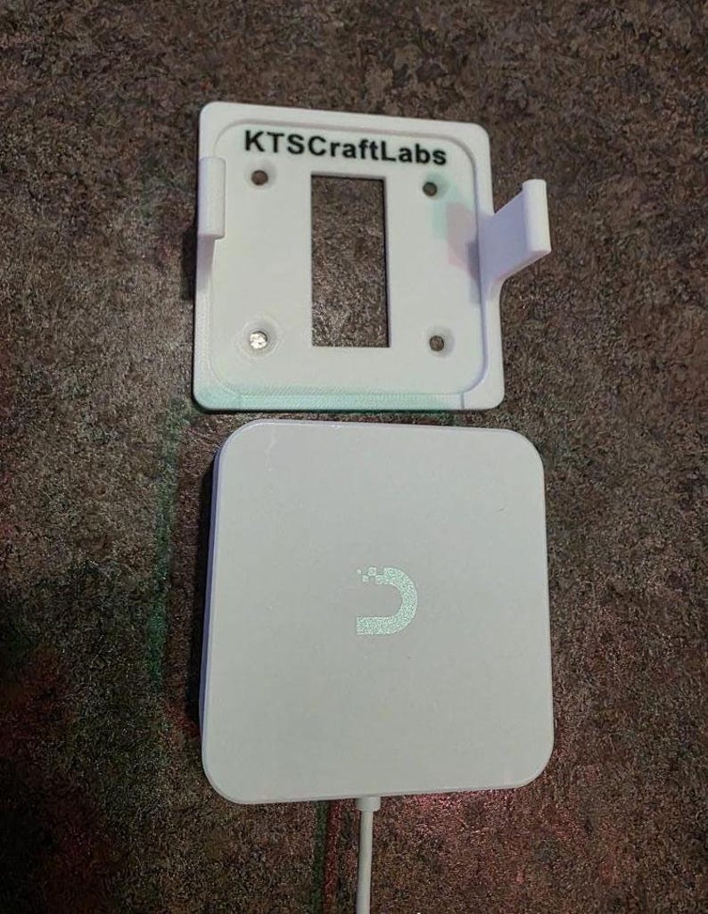 May include: A white wall mount with the text "KTSCraftLabs" above a rectangular aperture, and a white, square wireless charger with a curved design. A white cable extends from the charger.