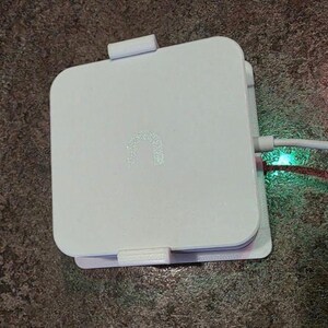 May include: A white, square electronic device with rounded corners. The device has a small, illuminated logo and is connected to a white cable. The cable is plugged in, and a green light is visible.