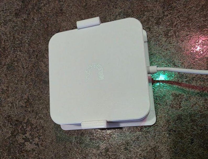 May include: A white, square electronic device with rounded corners. The device has a small, illuminated logo and is connected to a white cable. The cable is plugged in, and a green light is visible.