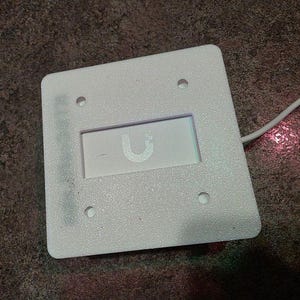 May include: A white, square electronic device with rounded corners. It has a recessed rectangular area with a stylized "U" logo. Four small holes are visible, and a white cable extends from the device.