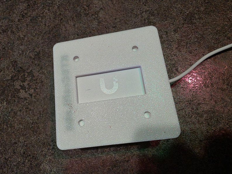 May include: A white, square electronic device with rounded corners. It has a recessed rectangular area with a stylized "U" logo. Four small holes are visible, and a white cable extends from the device.