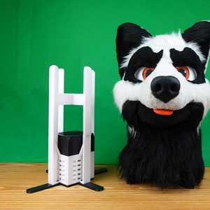 Pode incluir: Uma cabeça de animal peluda branca e preta com olhos laranja e um nariz preto. A cabeça está sobre uma superfície de madeira ao lado de um suporte branco com pernas pretas.