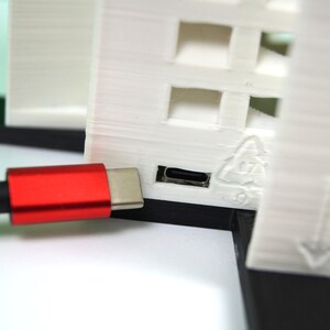 K&ouml;nnte beinhalten: Ein rotes USB-C-Kabel, das in ein wei&szlig;es 3D-gedrucktes Ger&auml;t mit einem Recycling-Symbol und einem nach unten zeigenden Pfeil gesteckt ist.