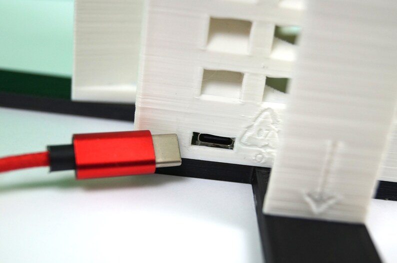 K&ouml;nnte beinhalten: Ein rotes USB-C-Kabel, das in ein wei&szlig;es 3D-gedrucktes Ger&auml;t mit einem Recycling-Symbol und einem nach unten zeigenden Pfeil gesteckt ist.