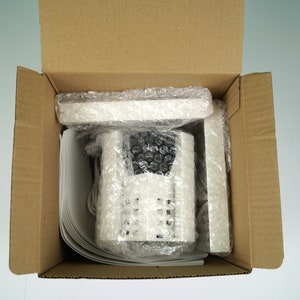 K&ouml;nnte beinhalten: Eine wei&szlig;e Sicherheitskamera mit einem schwarzen Objektiv ist in einem braunen Karton mit Luftpolsterfolie verpackt.