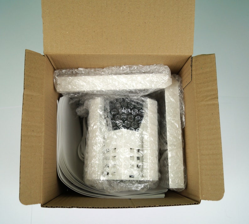 K&ouml;nnte beinhalten: Eine wei&szlig;e Sicherheitskamera mit einem schwarzen Objektiv ist in einem braunen Karton mit Luftpolsterfolie verpackt.