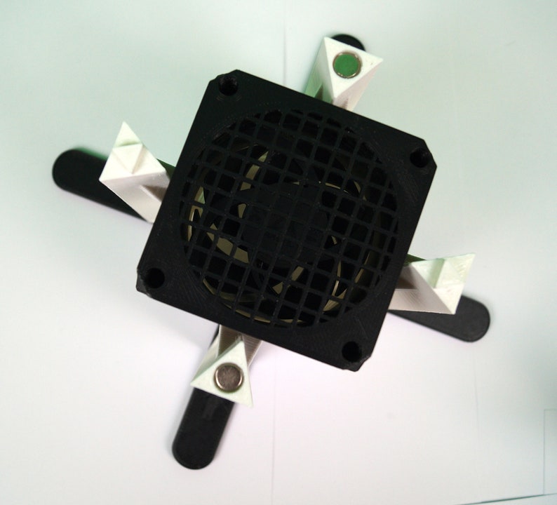 K&ouml;nnte beinhalten: Ein schwarzer Computerl&uuml;fter, der auf einem wei&szlig;en Kunststoffst&auml;nder mit vier Beinen montiert ist. Der L&uuml;fter hat einen schwarzen Kunststoffrahmen und ein schwarzes Metallgitter.