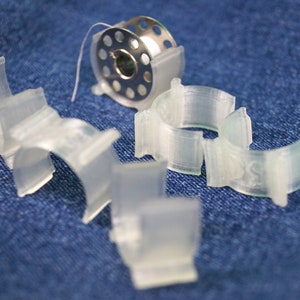 Puede incluir: Un juego de seis bobinas de plástico transparente para una máquina de coser. Las bobinas están diseñadas para sujetar hilo y están dispuestas sobre una superficie de mezclilla azul.