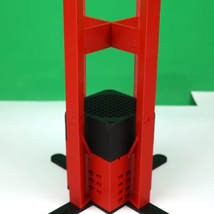 K&ouml;nnte beinhalten: Ein roter 3D-gedruckter St&auml;nder mit schwarzem Sockel und vier Beinen. Der St&auml;nder ist so konzipiert, dass er ein kleines Ger&auml;t oder Objekt h&auml;lt.