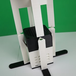 K&ouml;nnte beinhalten: Ein wei&szlig;er 3D-gedruckter St&auml;nder mit einem schwarzen quadratischen Objekt in der Mitte. Der St&auml;nder hat vier Beine und ist so konzipiert, dass er das Objekt an seinem Platz h&auml;lt.