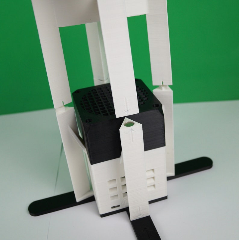 K&ouml;nnte beinhalten: Ein wei&szlig;er 3D-gedruckter St&auml;nder mit einem schwarzen quadratischen Objekt in der Mitte. Der St&auml;nder hat vier Beine und ist so konzipiert, dass er das Objekt an seinem Platz h&auml;lt.