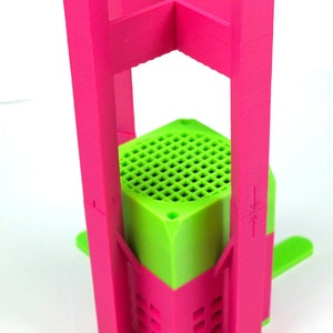 K&ouml;nnte beinhalten: Ein rosa und gr&uuml;ner 3D-gedruckter St&auml;nder mit sechseckiger Basis und vier hohen Beinen. Der St&auml;nder ist so konzipiert, dass er einen kleinen Gegenstand wie einen Lautsprecher oder eine Pflanze aufnehmen kann.