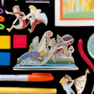 May include: Enamel pin featuring a futuristic city design with a rocket ship taking off. The pin has a blue, white, red, and yellow color scheme. Other pins and tools are visible in the background.